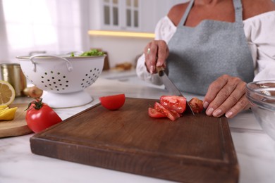 Photo of Senior woman cutting tomato at white marble table in kitchen, closeup