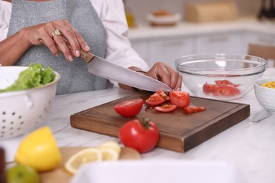 Photo of Senior woman cutting tomato at white marble table in kitchen, closeup