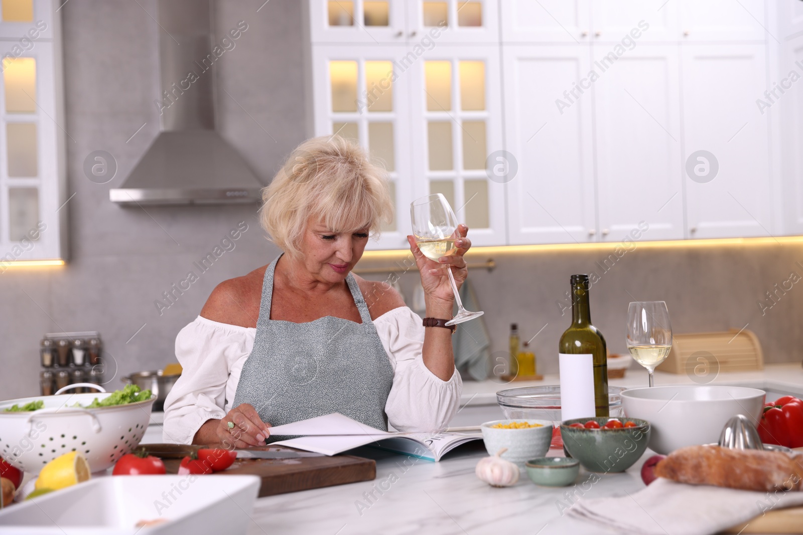 Senior woman reading magazine and drinking white wine at table in kitchen Photo of Senior woman reading magazine and drinking white wine at table in kitchen