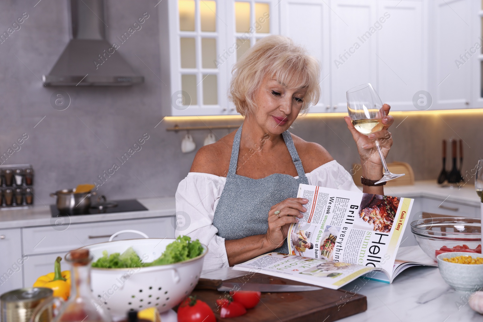 Senior woman reading magazine and drinking white wine at table in kitchen Photo of Senior woman reading magazine and drinking white wine at table in kitchen