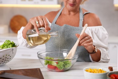 Photo of Senior woman pouring oil into bowl of salad at marble table indoors, closeup