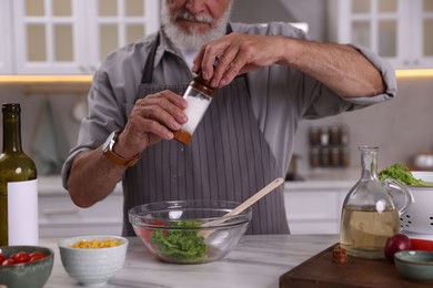 Photo of Elderly man salting salad at marble table in kitchen, closeup