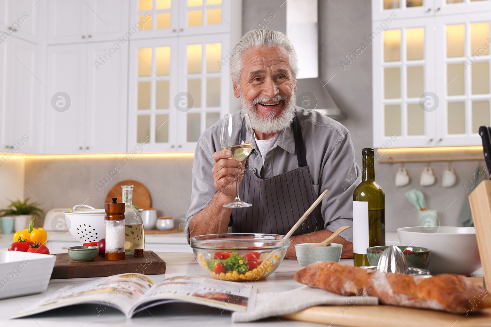 Smiling elderly man drinking white wine after cooking at table in kitchen Photo of Smiling elderly man drinking white wine after cooking at table in kitchen