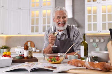 Photo of Smiling elderly man drinking white wine after cooking at table in kitchen