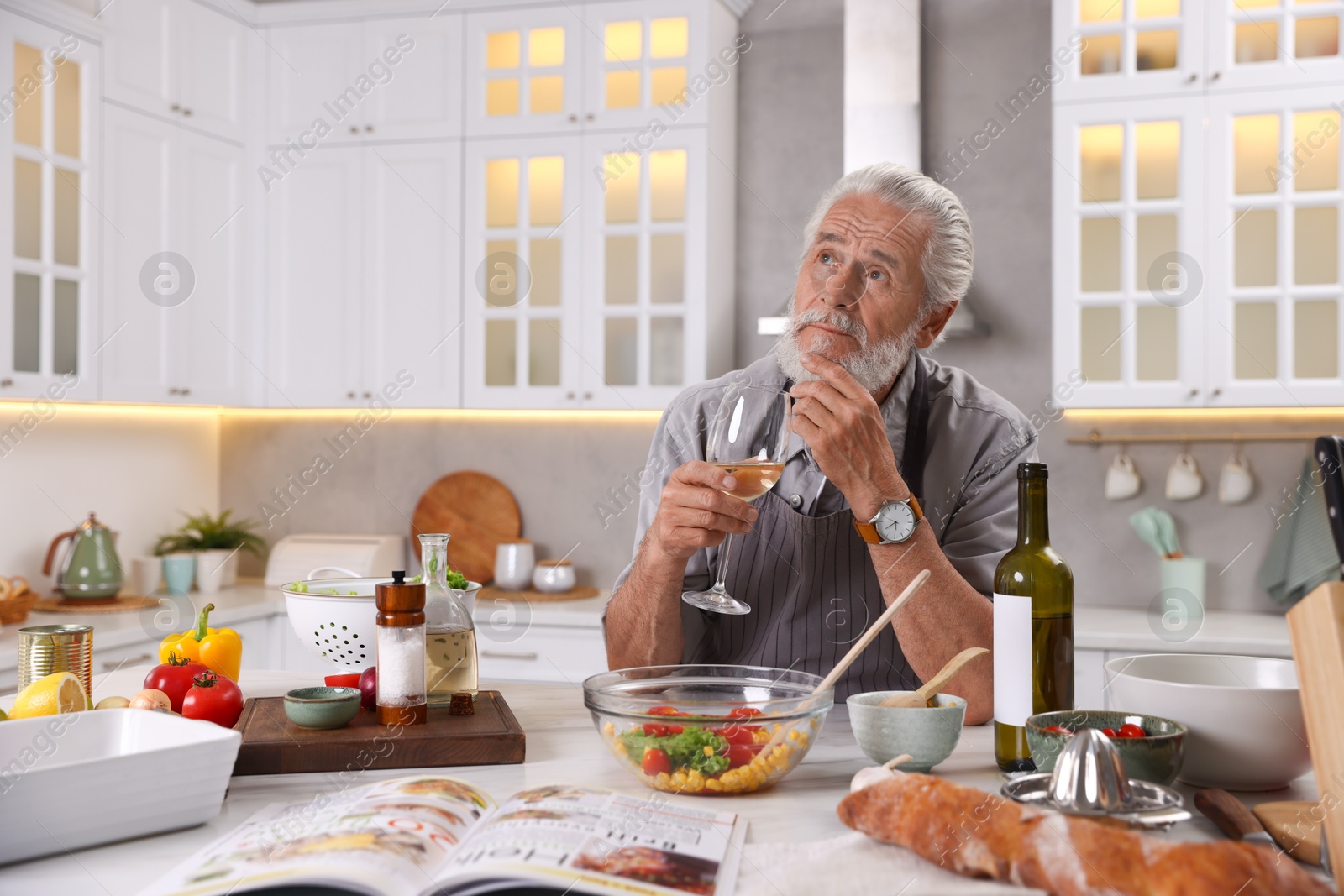 Thoughtful elderly man drinking white wine after cooking at table in kitchen Photo of Thoughtful elderly man drinking white wine after cooking at table in kitchen
