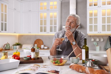 Photo of Thoughtful elderly man drinking white wine after cooking at table in kitchen