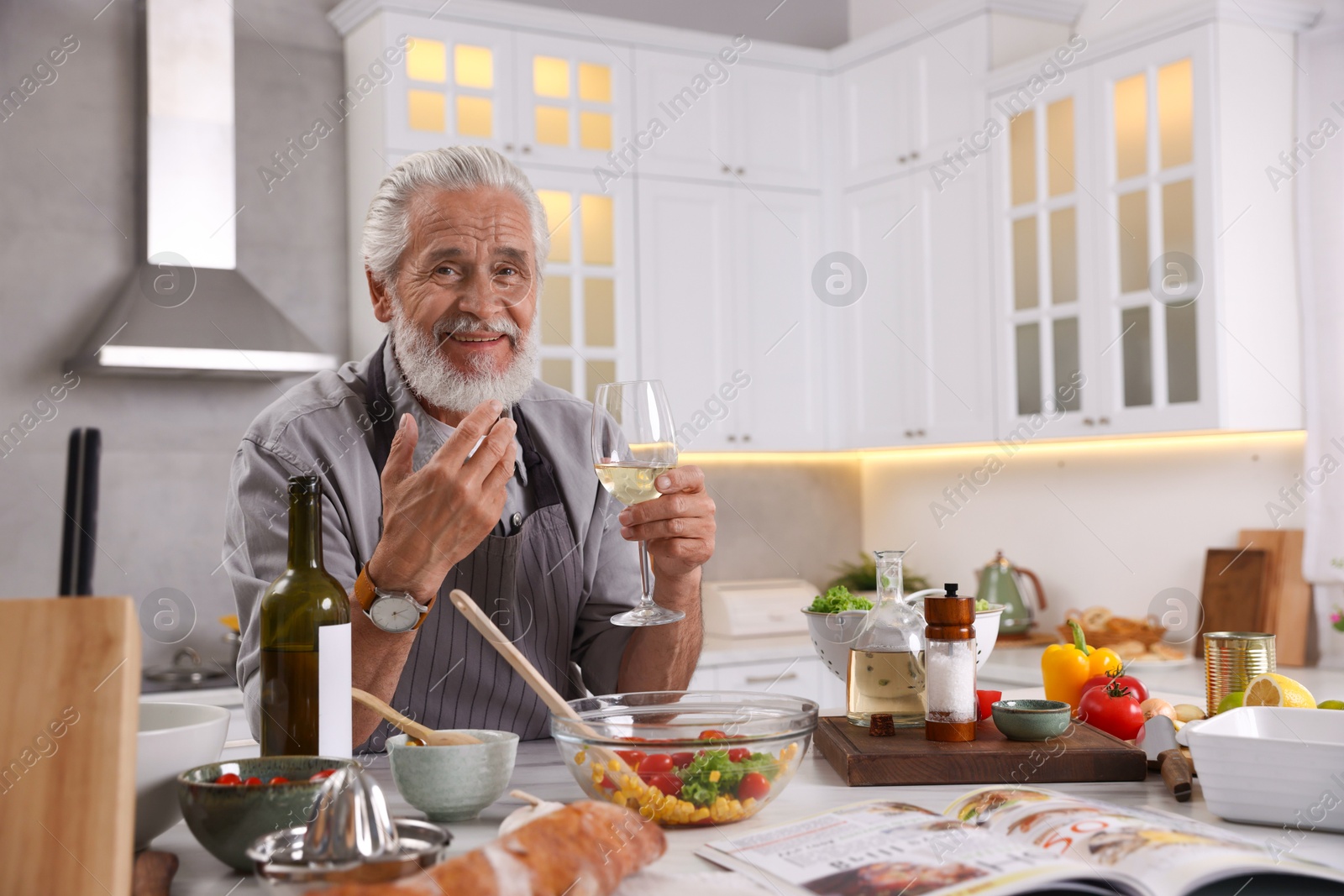 Smiling elderly man drinking white wine after cooking at table in kitchen Photo of Smiling elderly man drinking white wine after cooking at table in kitchen