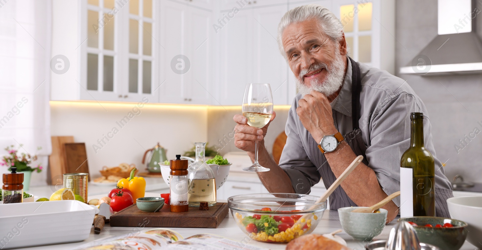 Smiling elderly man drinking white wine after cooking at table in kitchen Photo of Smiling elderly man drinking white wine after cooking at table in kitchen