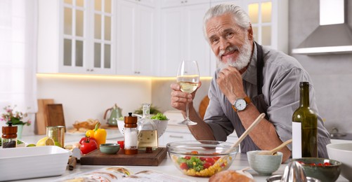 Photo of Smiling elderly man drinking white wine after cooking at table in kitchen