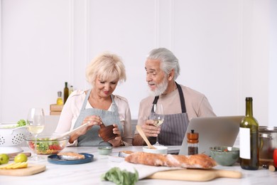 Photo of Elderly couple with laptop cooking together and drinking white wine at table indoors