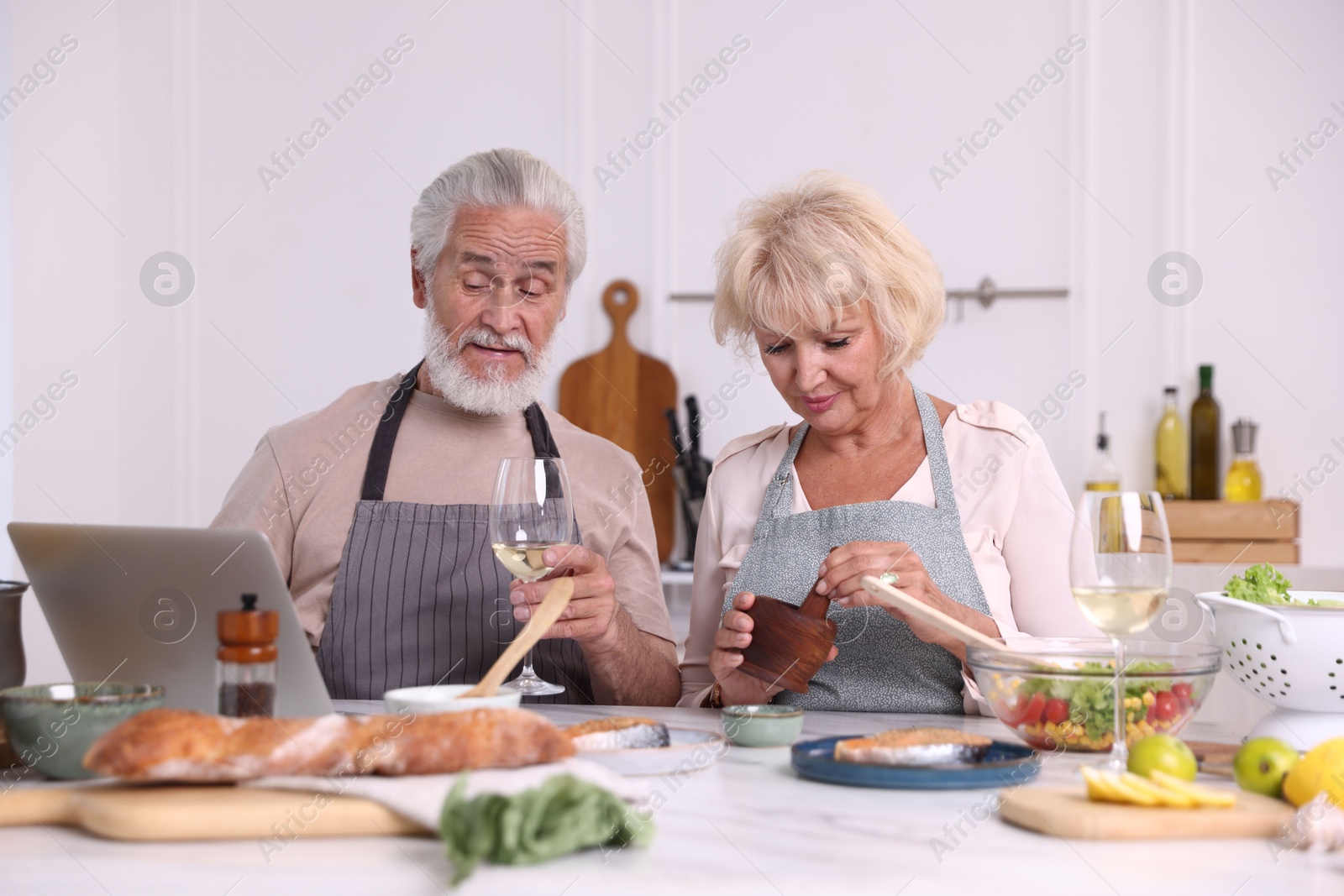 Elderly couple with laptop cooking together and drinking white wine at table indoors Photo of Elderly couple with laptop cooking together and drinking white wine at table indoors
