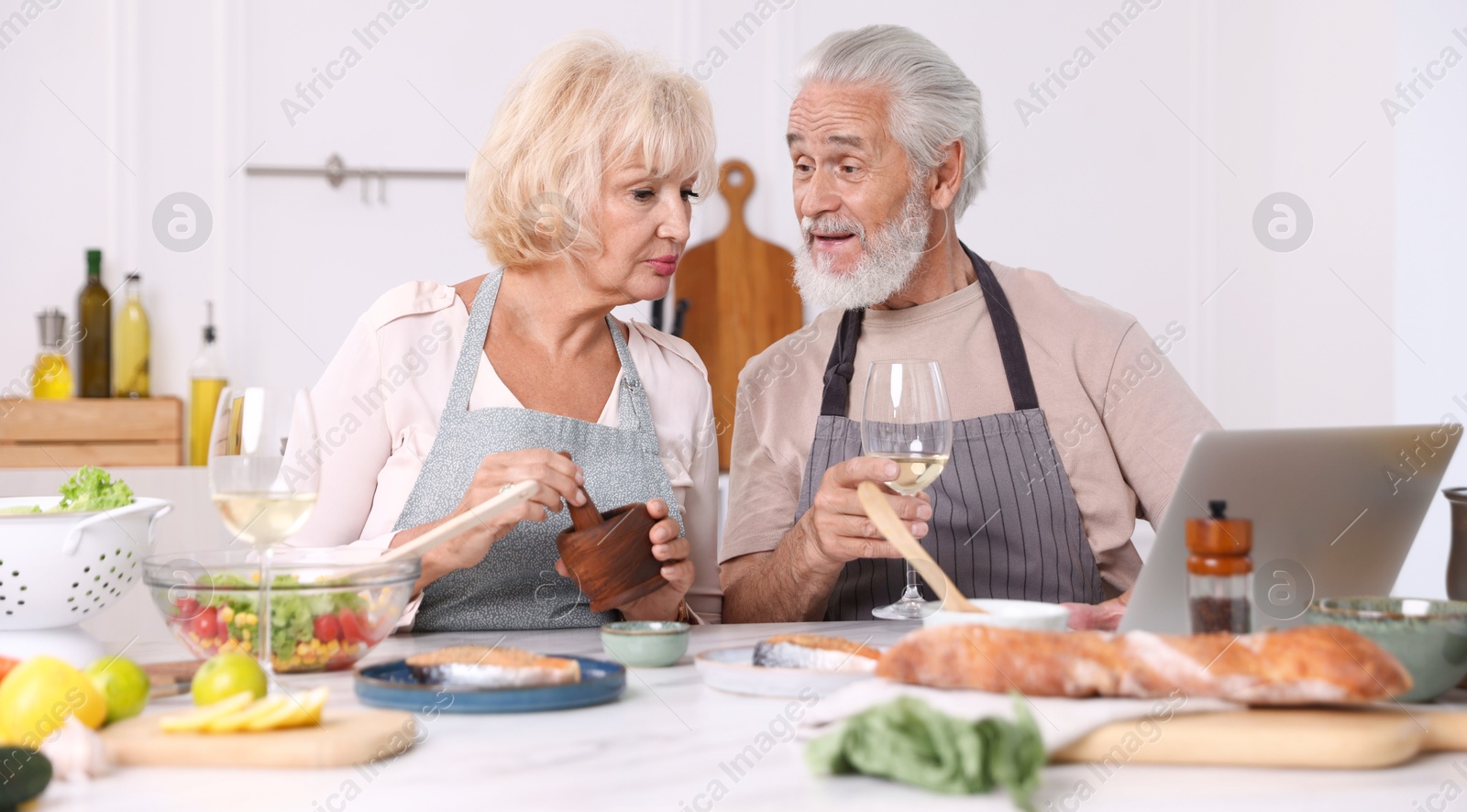 Elderly couple with laptop cooking together and drinking white wine at table indoors Photo of Elderly couple with laptop cooking together and drinking white wine at table indoors