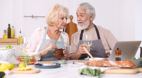 Photo of Elderly couple with laptop cooking together and drinking white wine at table indoors