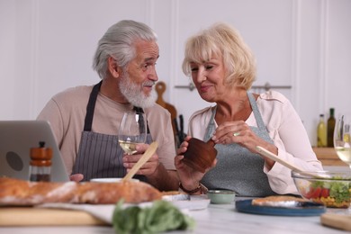 Photo of Elderly couple with laptop cooking together and drinking white wine at table indoors