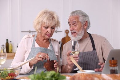 Photo of Elderly couple cooking together and drinking white wine at table indoors