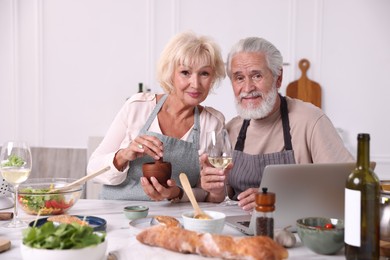 Photo of Portrait of elderly couple with laptop cooking together and drinking white wine at table indoors