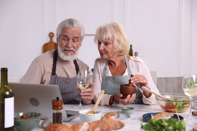 Photo of Elderly couple with laptop cooking together and drinking white wine at table indoors
