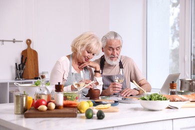 Photo of Elderly couple cooking together and drinking white wine at counter indoors