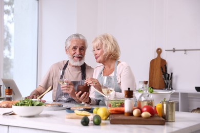 Photo of Elderly couple cooking together and drinking white wine at counter indoors