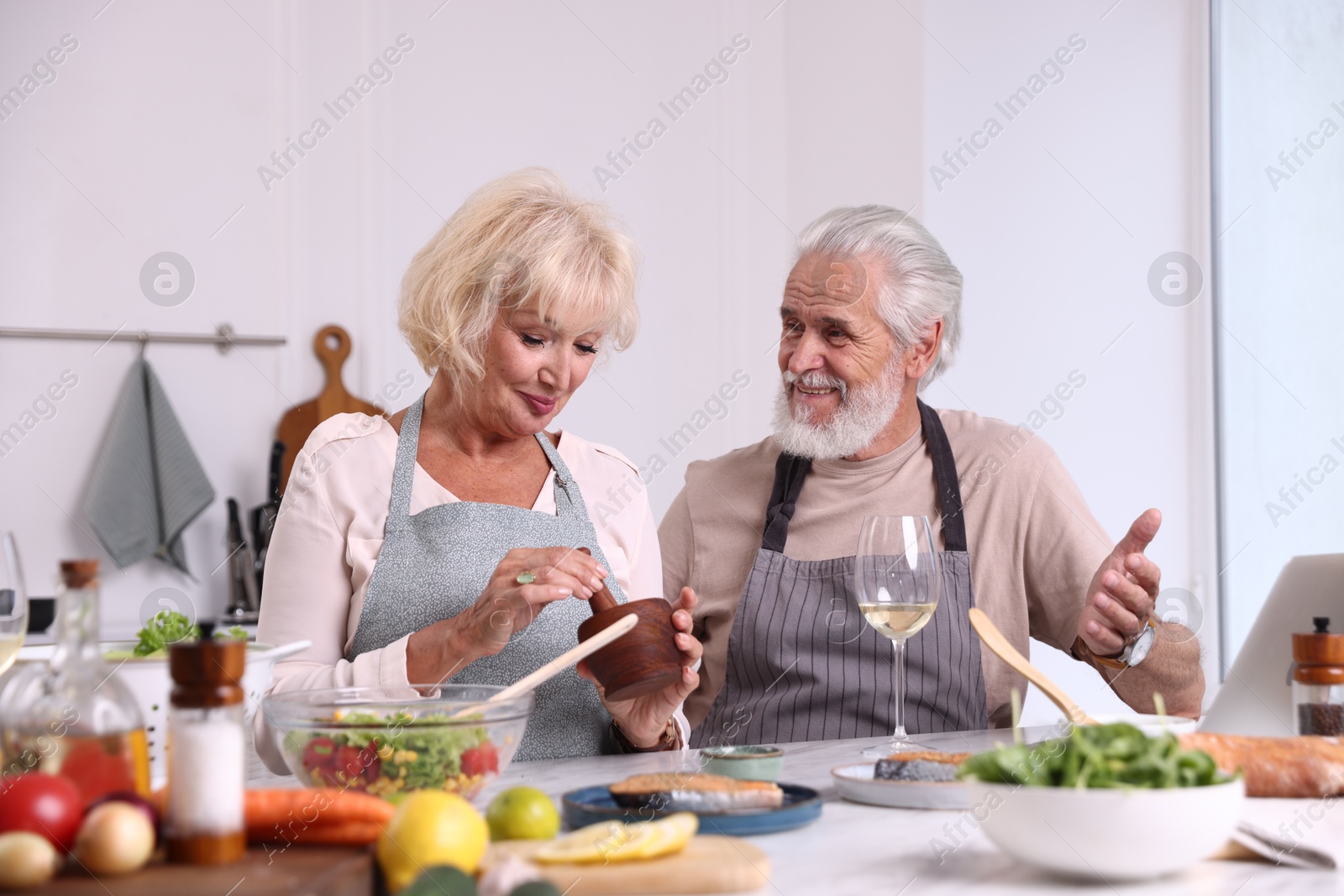 Elderly couple cooking together and drinking white wine at table indoors Photo of Elderly couple cooking together and drinking white wine at table indoors
