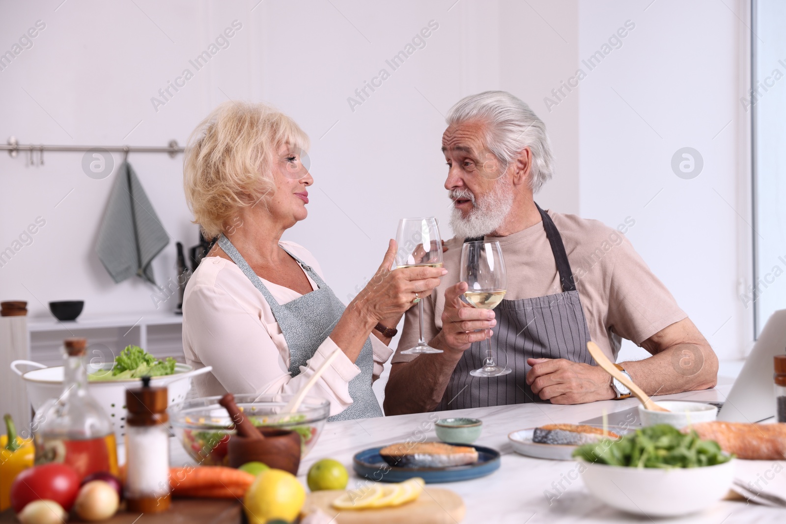 Elderly couple drinking white wine while cooking at table in kitchen Photo of Elderly couple drinking white wine while cooking at table in kitchen