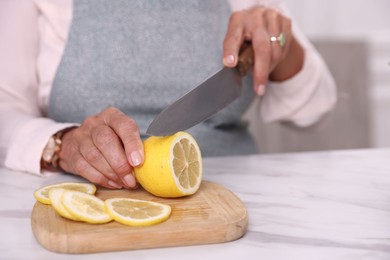 Photo of Senior woman cutting lemon at white marble table indoors, closeup