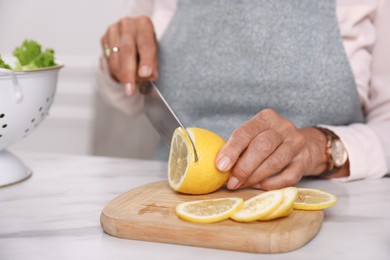 Photo of Senior woman cutting lemon at white marble table indoors, closeup