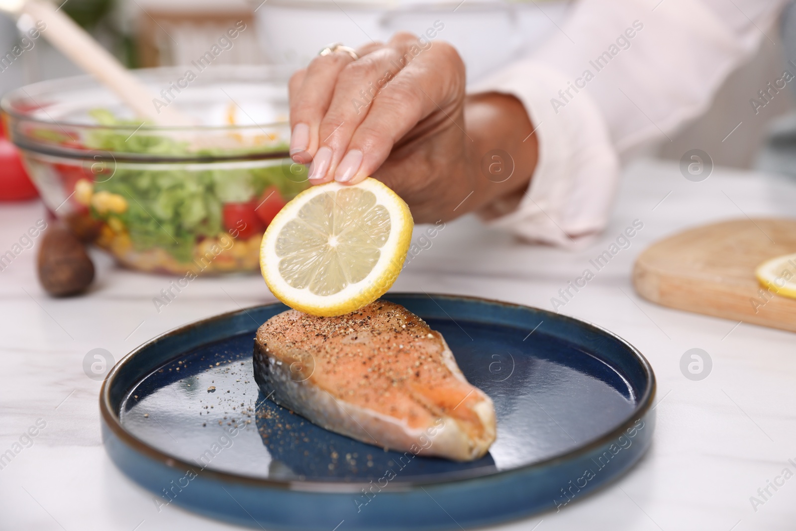 Senior woman putting slice of lemon onto salmon fish at white marble table indoors, closeup Photo of Senior woman putting slice of lemon onto salmon fish at white marble table indoors, closeup