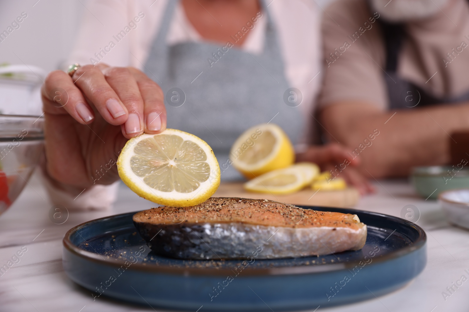 Senior woman putting slice of lemon onto salmon fish at white marble table indoors, closeup Photo of Senior woman putting slice of lemon onto salmon fish at white marble table indoors, closeup