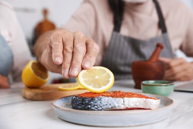 Photo of Elderly man putting slice of lemon onto salmon fish at white marble table indoors, closeup