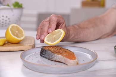 Photo of Elderly man putting slice of lemon onto salmon fish at white marble table indoors, closeup