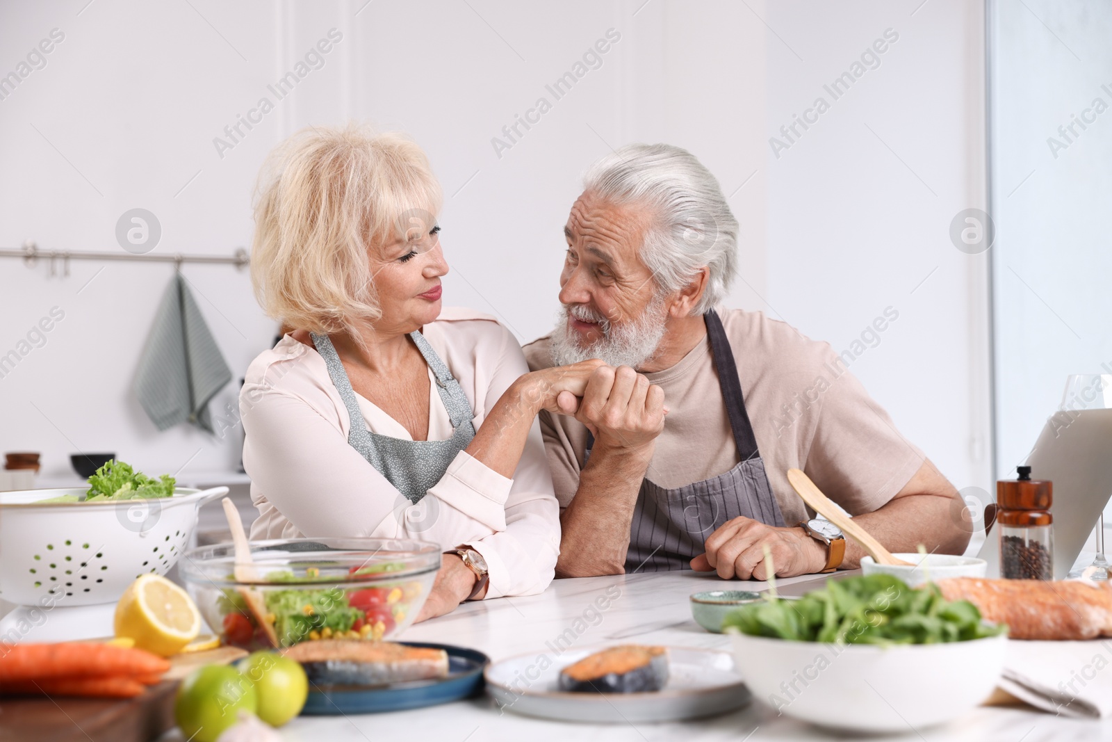 Elderly couple holding hands while cooking together at table indoors Photo of Elderly couple holding hands while cooking together at table indoors