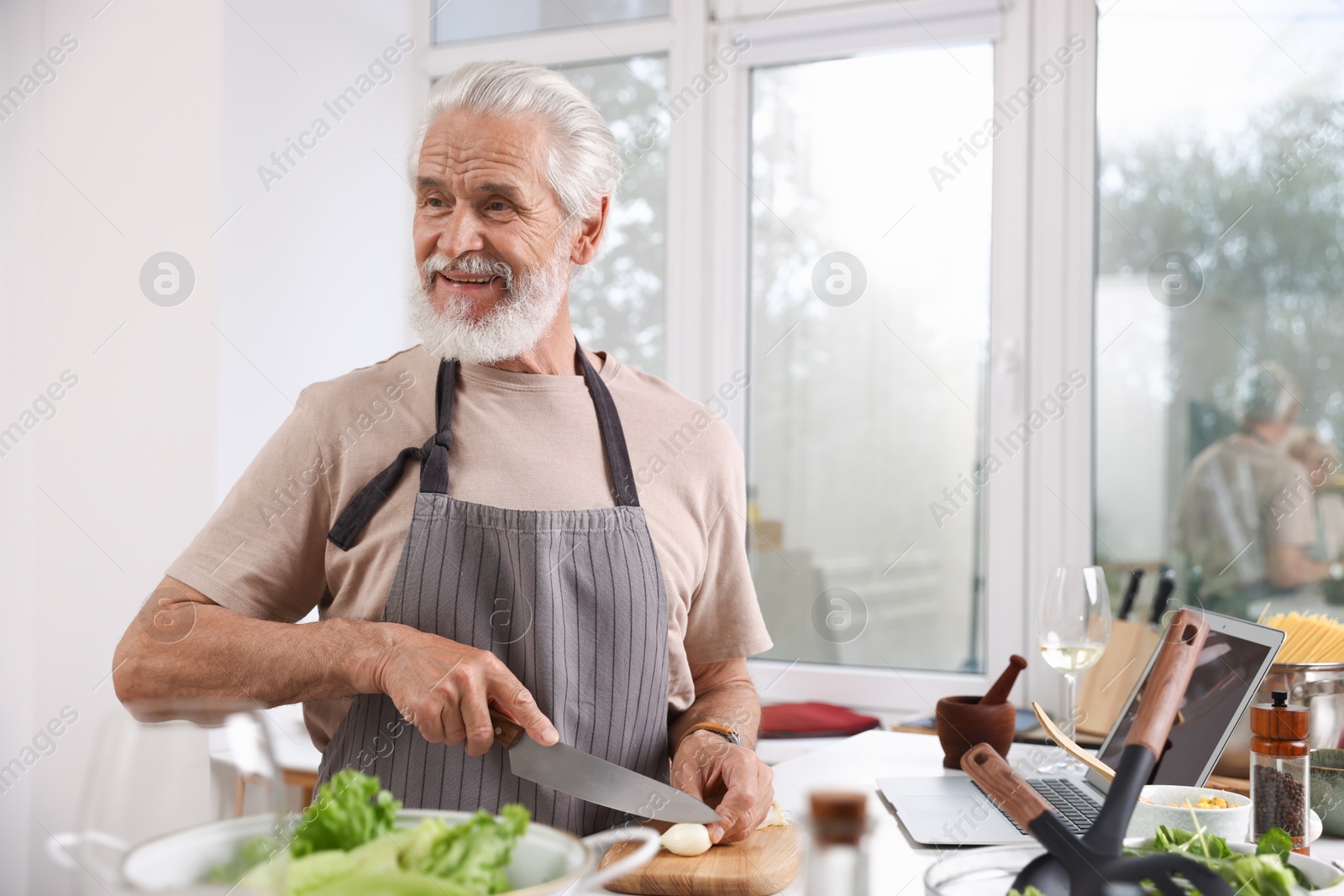 Smiling elderly man cooking at table near window indoors Photo of Smiling elderly man cooking at table near window indoors