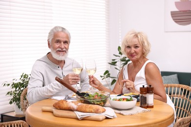 Photo of Elderly couple clinking glasses of white wine during dinner at table indoors