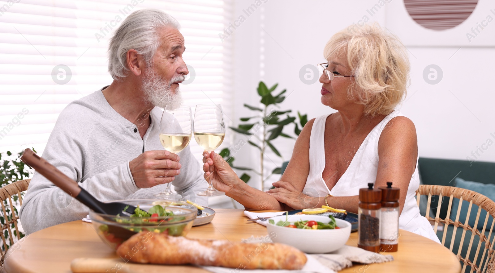 Elderly couple clinking glasses of white wine during dinner at table indoors Photo of Elderly couple clinking glasses of white wine during dinner at table indoors