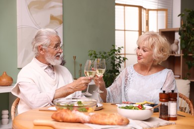 Photo of Elderly couple clinking glasses of white wine during dinner at table indoors