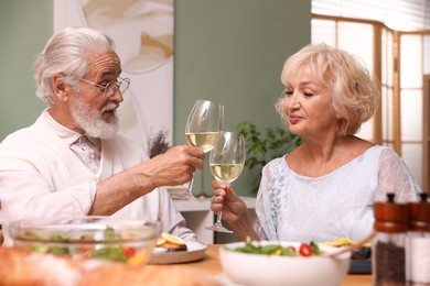 Photo of Elderly couple clinking glasses of white wine during dinner at table indoors