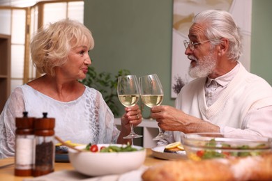 Photo of Elderly couple clinking glasses of white wine during dinner at table indoors