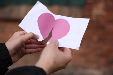 Image of Betrayal. Man tearing sheet of paper with pink heart into halves outdoors, closeup