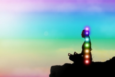 Image of Woman meditating in Lotus pose against rainbow sky. Seven chakras glowing