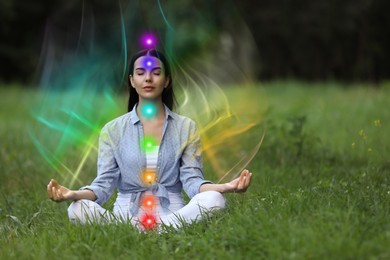 Image of Woman meditating in Lotus pose on green grass. Aura seen as rainbow waves around her. Seven chakras glowing