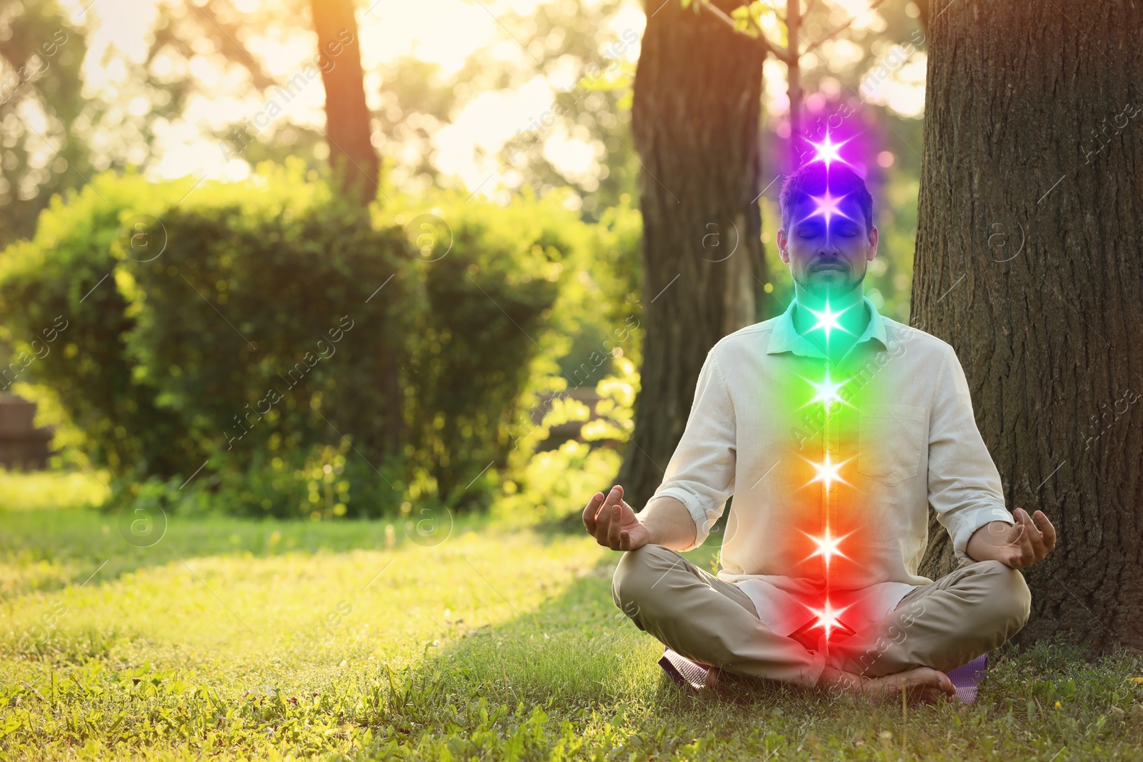 Image of Woman meditating in Lotus pose on green grass next to tree. Seven chakras glowing