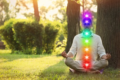 Image of Woman meditating in Lotus pose on green grass next to tree. Seven chakras glowing