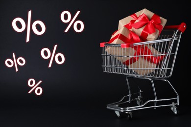 Image of Percent signs near shopping cart with gift boxes against black background. Hot sale, special offer