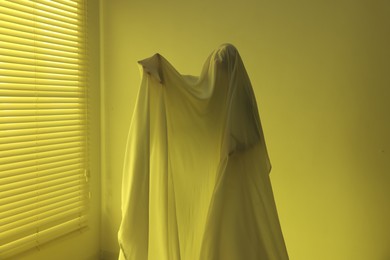 Photo of Creepy ghost. Man covered with white sheet indoors in yellow light