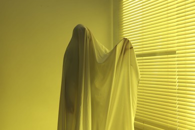 Photo of Creepy ghost. Man covered with white sheet indoors in yellow light
