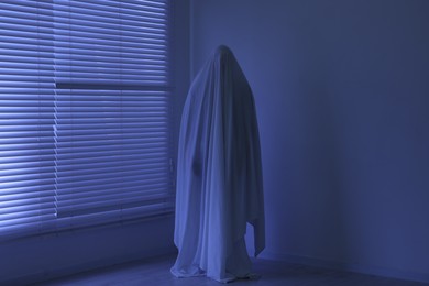 Photo of Creepy ghost. Man covered with white sheet indoors in blue light
