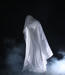 Photo of Creepy ghost. Man covered with white sheet in smoke on black background