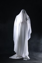 Photo of Creepy ghost. Man covered with white sheet in smoke on black background