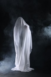 Photo of Creepy ghost. Man covered with white sheet in smoke on black background